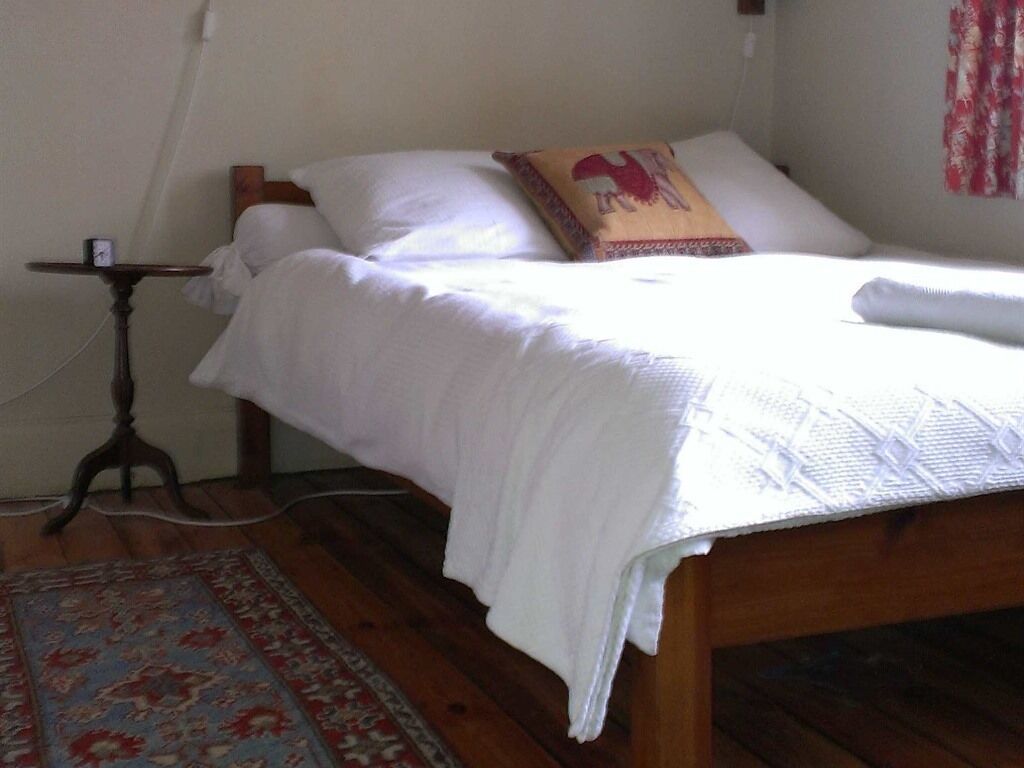 Una cama bien hecha con sábanas blancas y un cojín decorativo, una pequeña mesa redonda con un reloj y una alfombra estampada sobre un piso de madera.