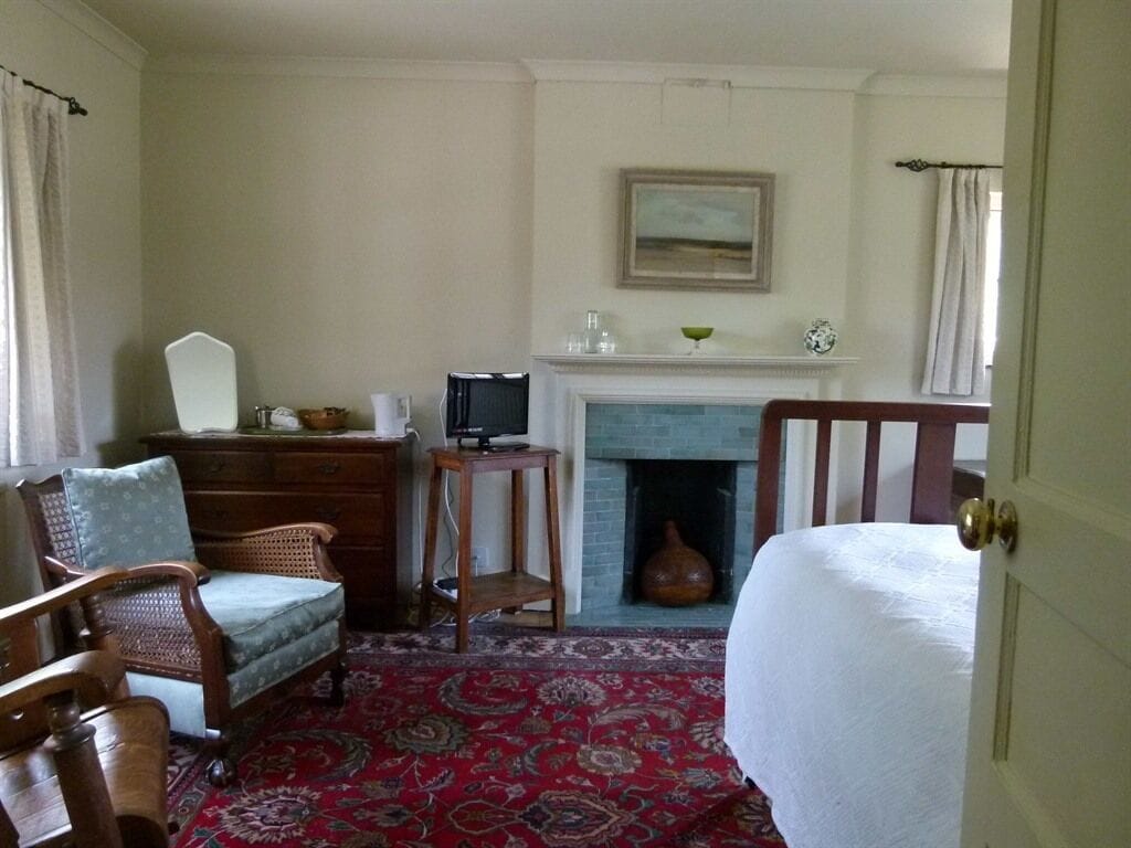 A room with a fireplace, a bed, a wooden chair, a small table, and a red patterned rug.