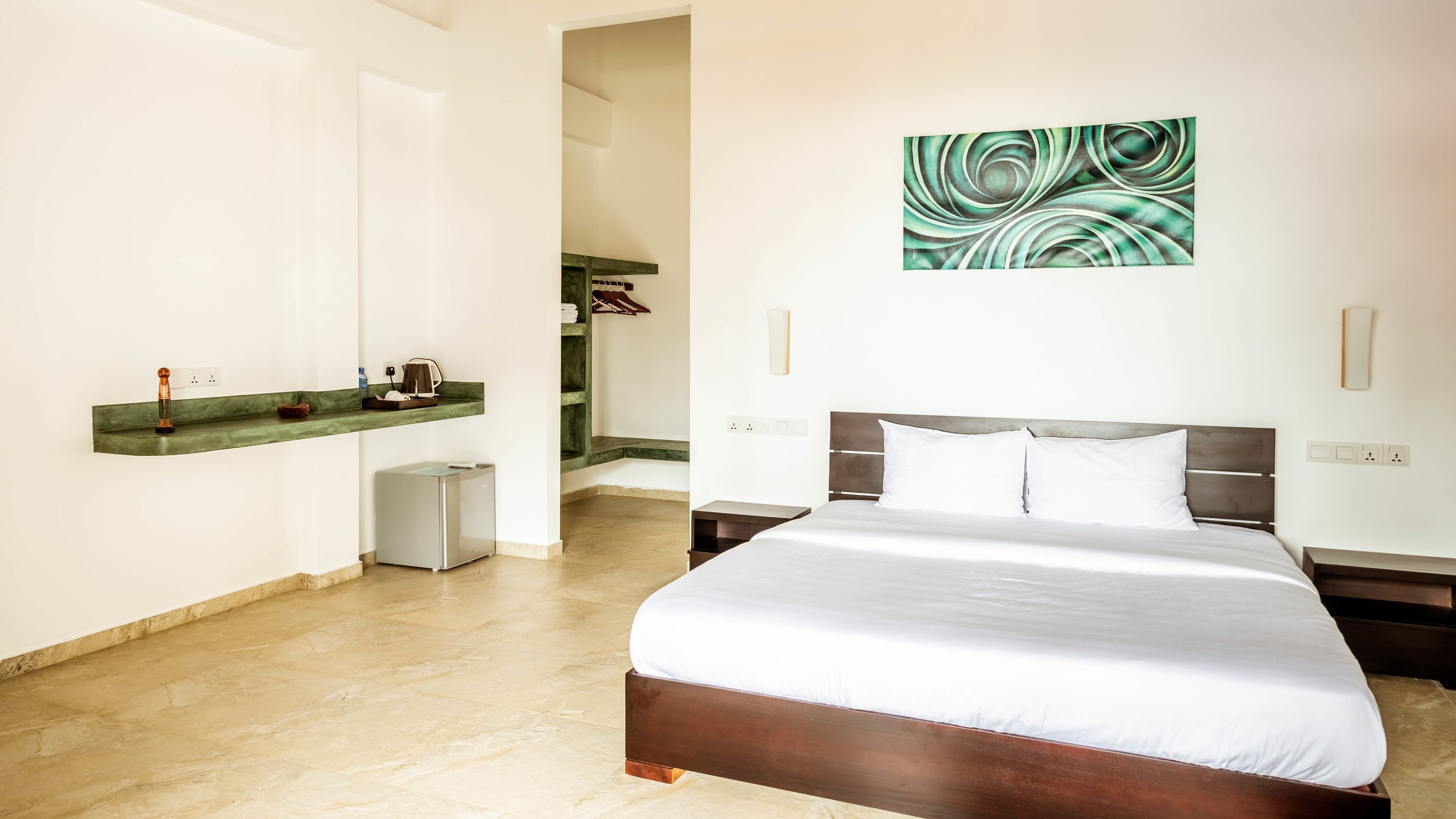 A hotel room with a large bed, a small bedside table, a wall-mounted artwork, and a built-in shelf with a kettle and a cup.
