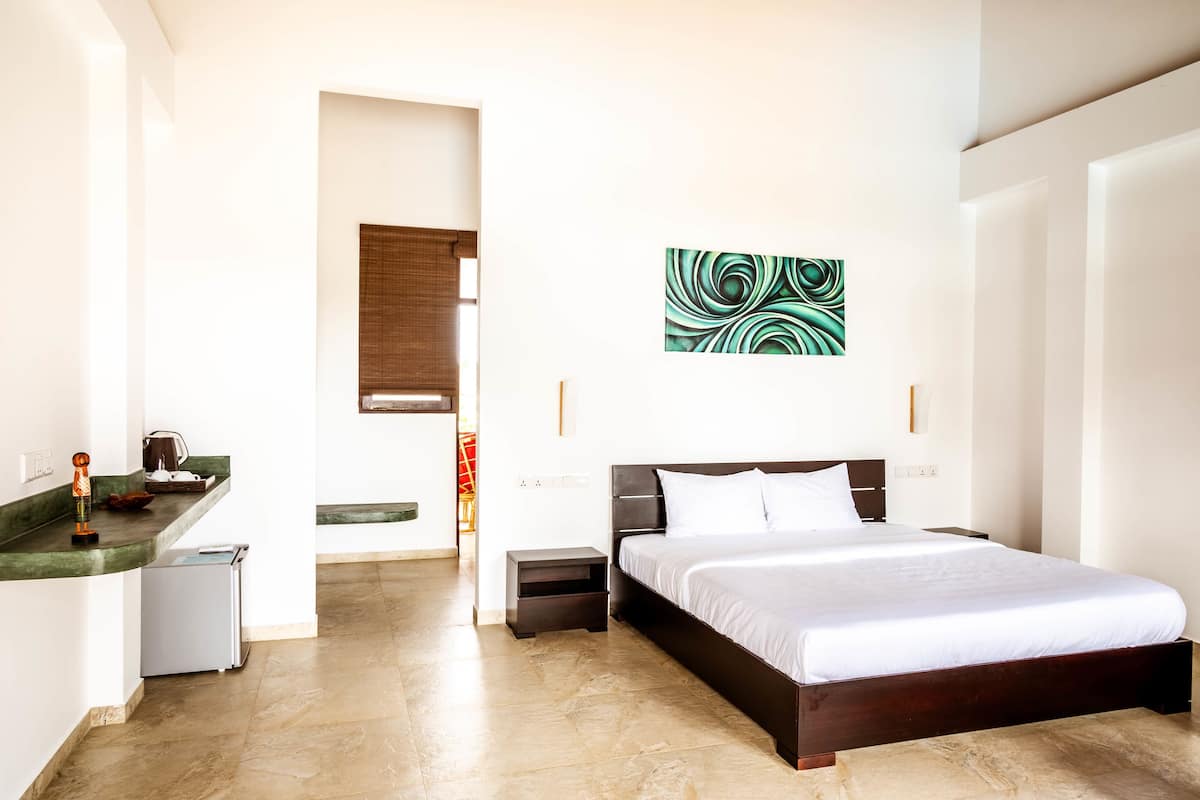 A modern hotel room with a large bed, a small bedside table, a nightstand, a built-in bench at the foot of the bed, a kitchen area with a counter and a refrigerator, and a wall-mounted abstract art piece.
