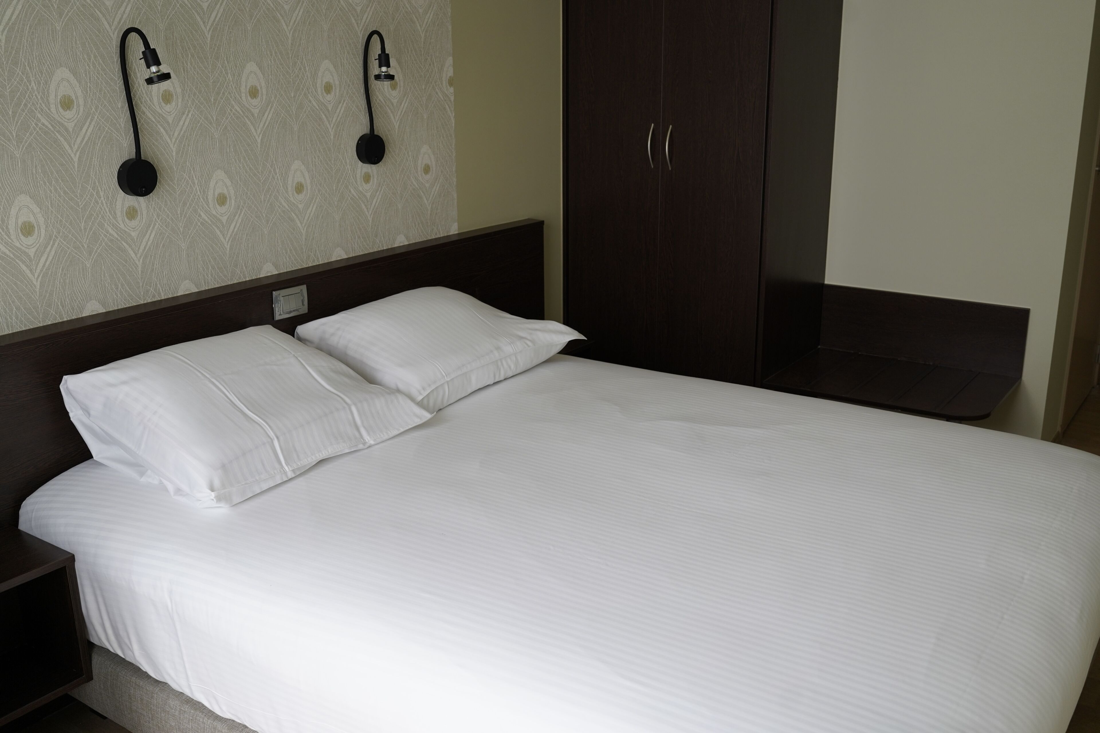 A neatly made bed with white linens and two pillows, a dark wooden headboard, and a wardrobe with two doors.