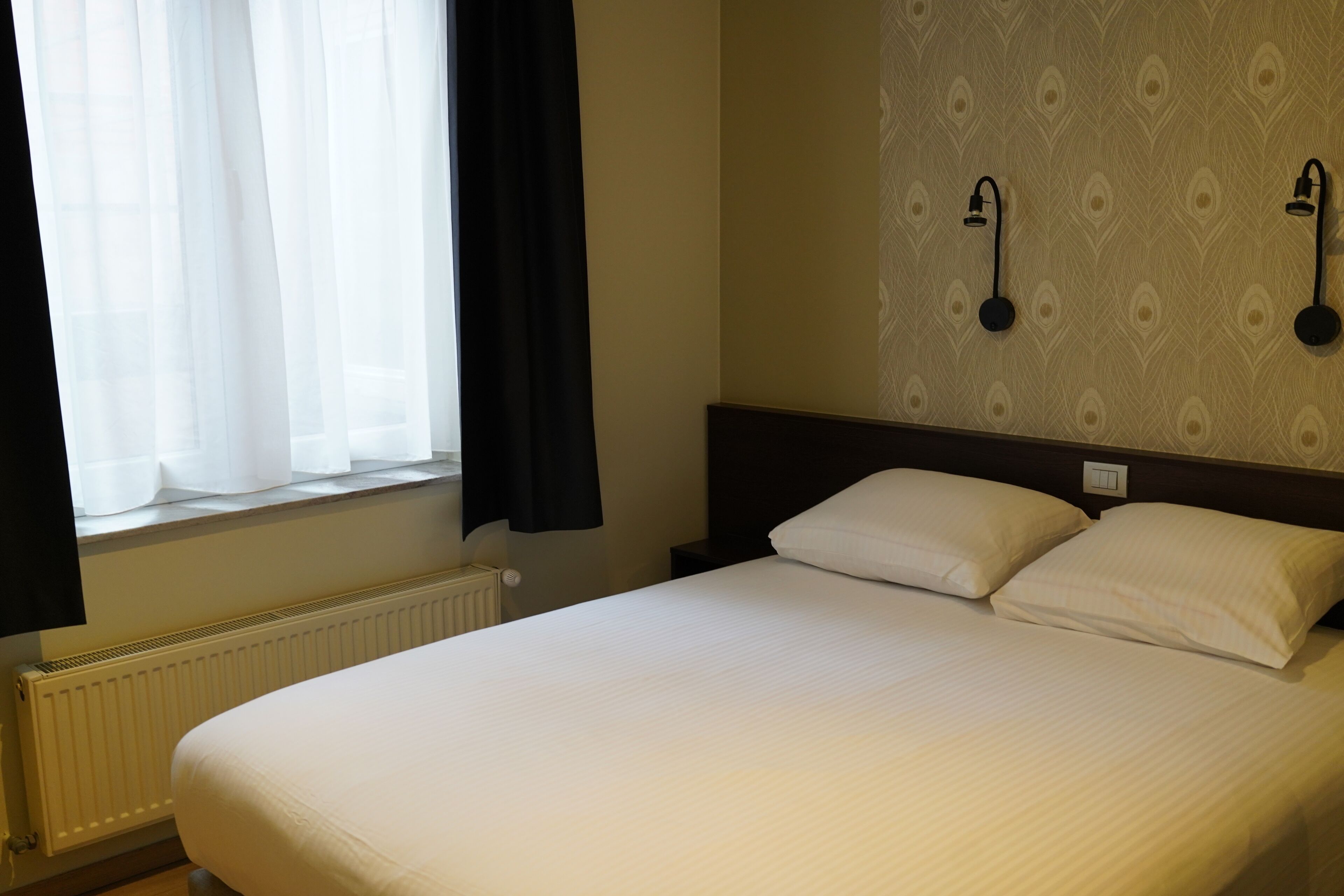 Habitación de hotel con una cama grande, una ventana con cortinas, un radiador y dos lámparas fijadas a la pared.
