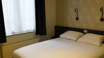 Habitación de hotel con una cama grande, una ventana con cortinas, un radiador y dos lámparas fijadas a la pared.