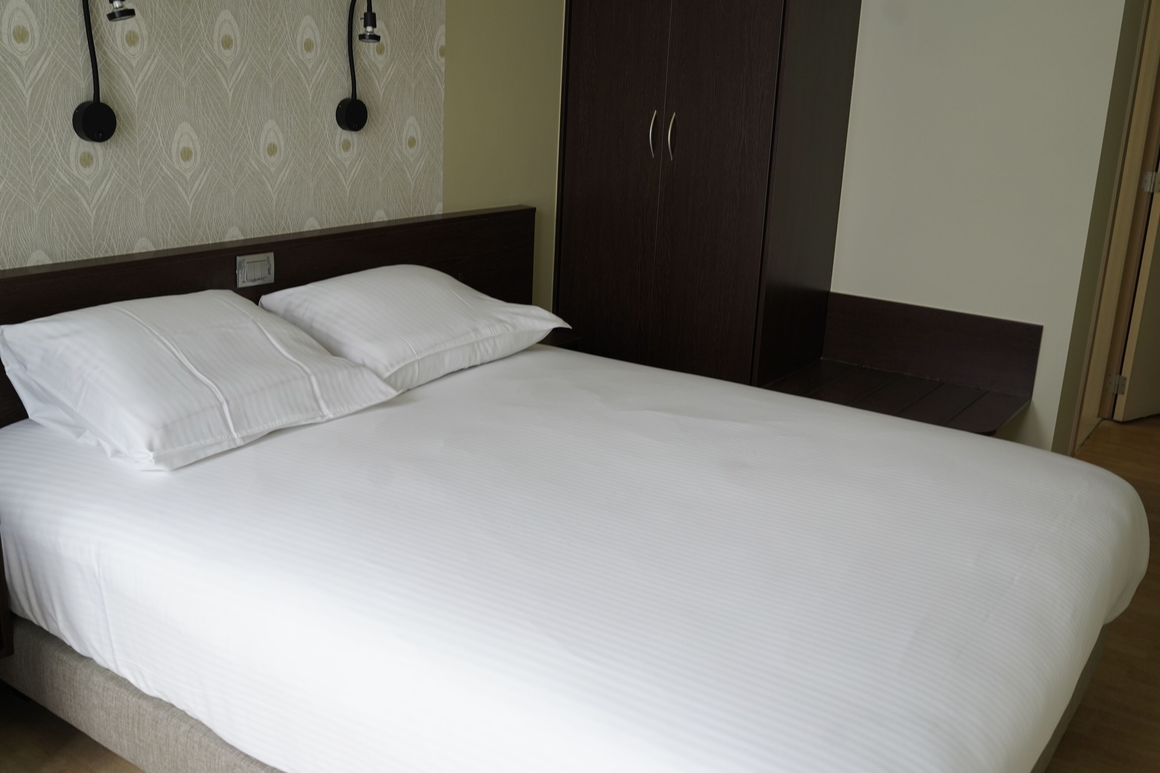 Una cama bien hecha con sábanas blancas y dos almohadas, un armario de madera oscura y una pared con diseño, con dos luces empotradas.