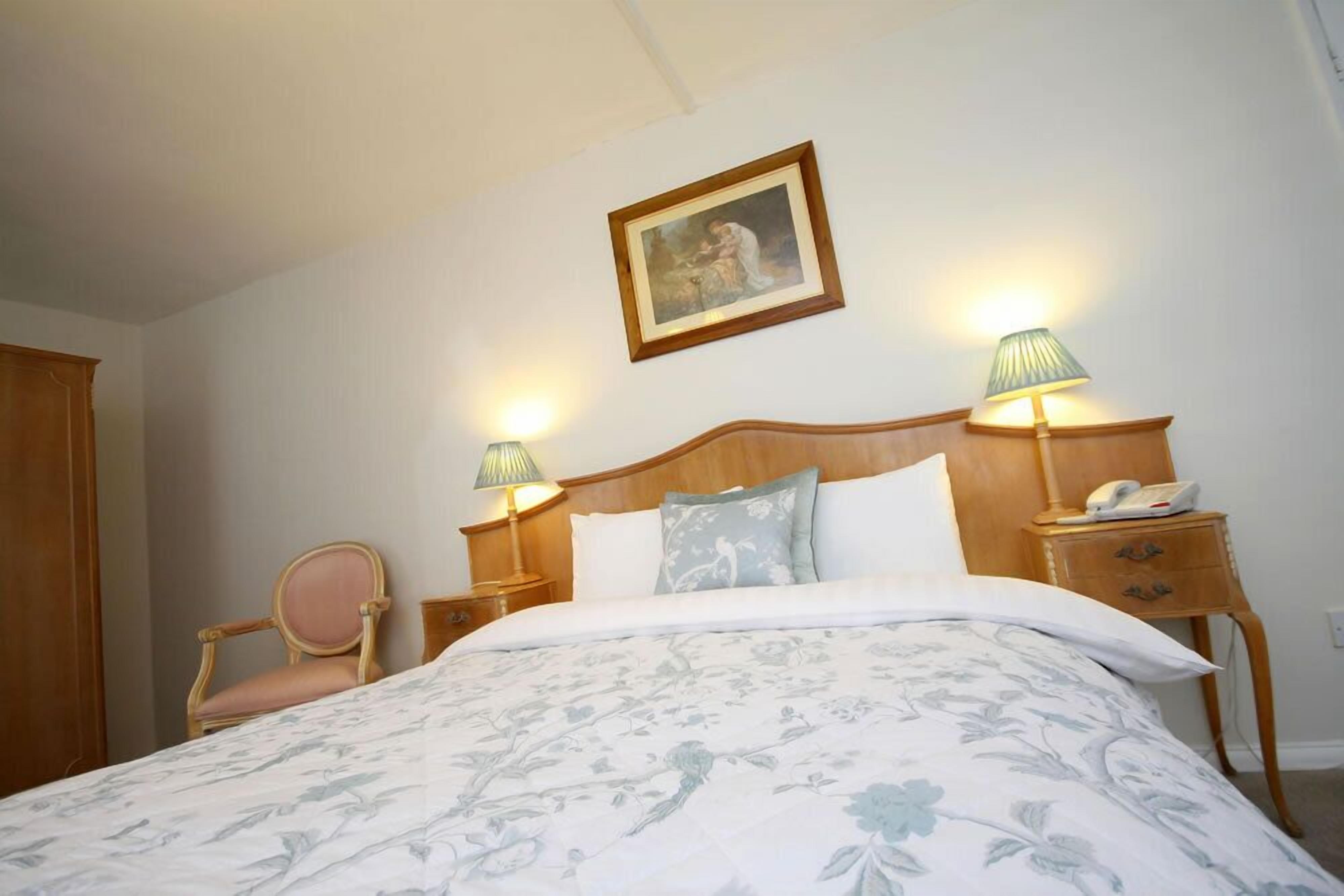 A neatly arranged bedroom with a large bed, two bedside lamps, a wooden nightstand, a chair, and a framed picture on the wall.