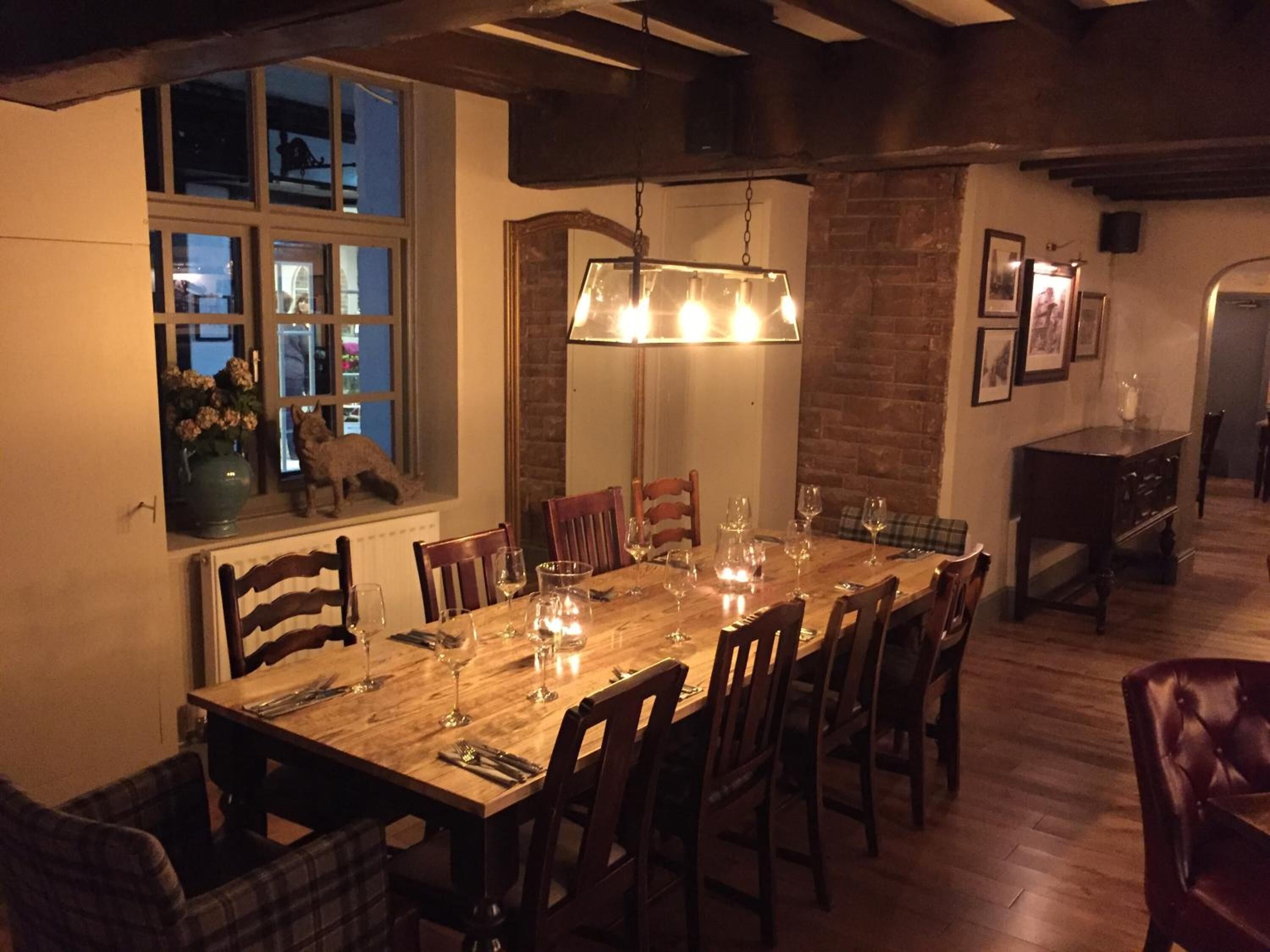 Un comedor con una larga mesa de madera dispuesta para seis personas, flanqueada por sillas de madera. Un lámpara colgante sobre la mesa proporciona iluminación.