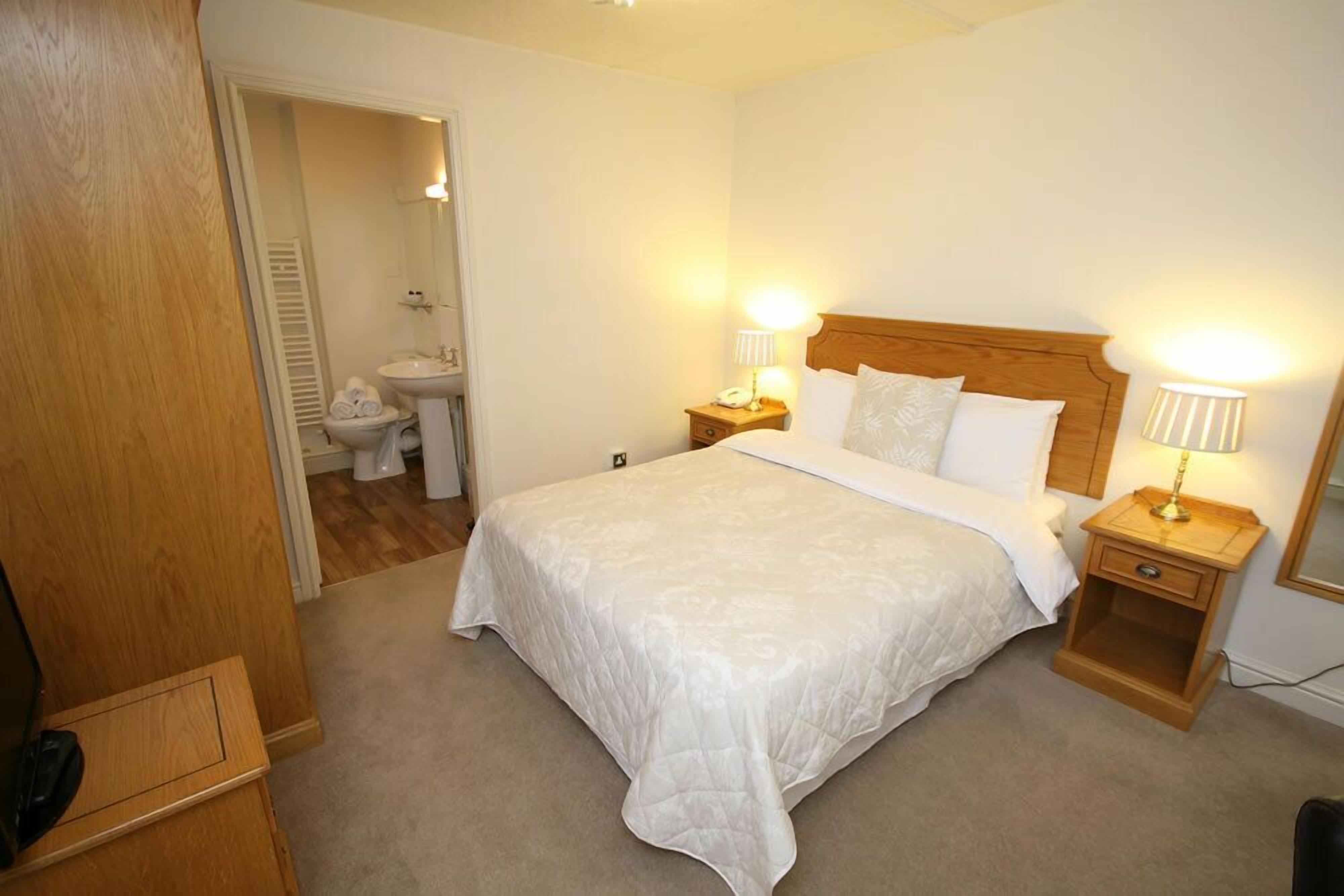 Una habitación de hotel con una cama grande, dos mesitas de noche con lámparas, un cabecero de madera, un armario de madera y un baño visible a través de una puerta abierta.