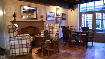 A cozy room with a fireplace, plaid chairs, a dining table with chairs, and a large window with a view of the street.