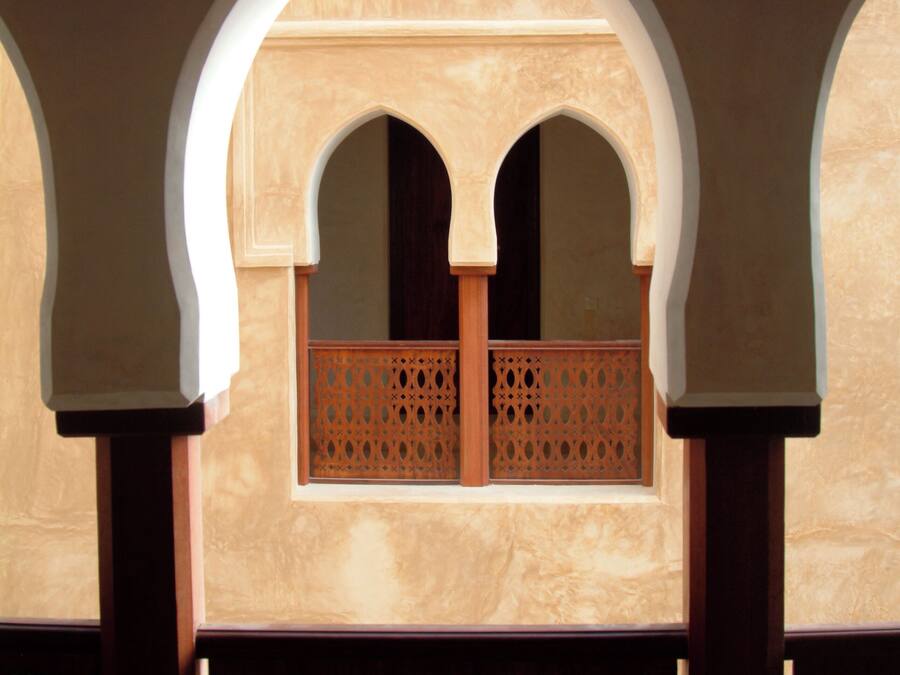 Ein rundbogenförmiges Fenster mit aufwendigem Holzgitter, eingelassen in eine beige Wand mit Marmoroptik.