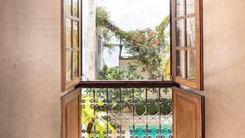 A view through an open window with a metal gate, leading to a balcony with plants and a view of a building.