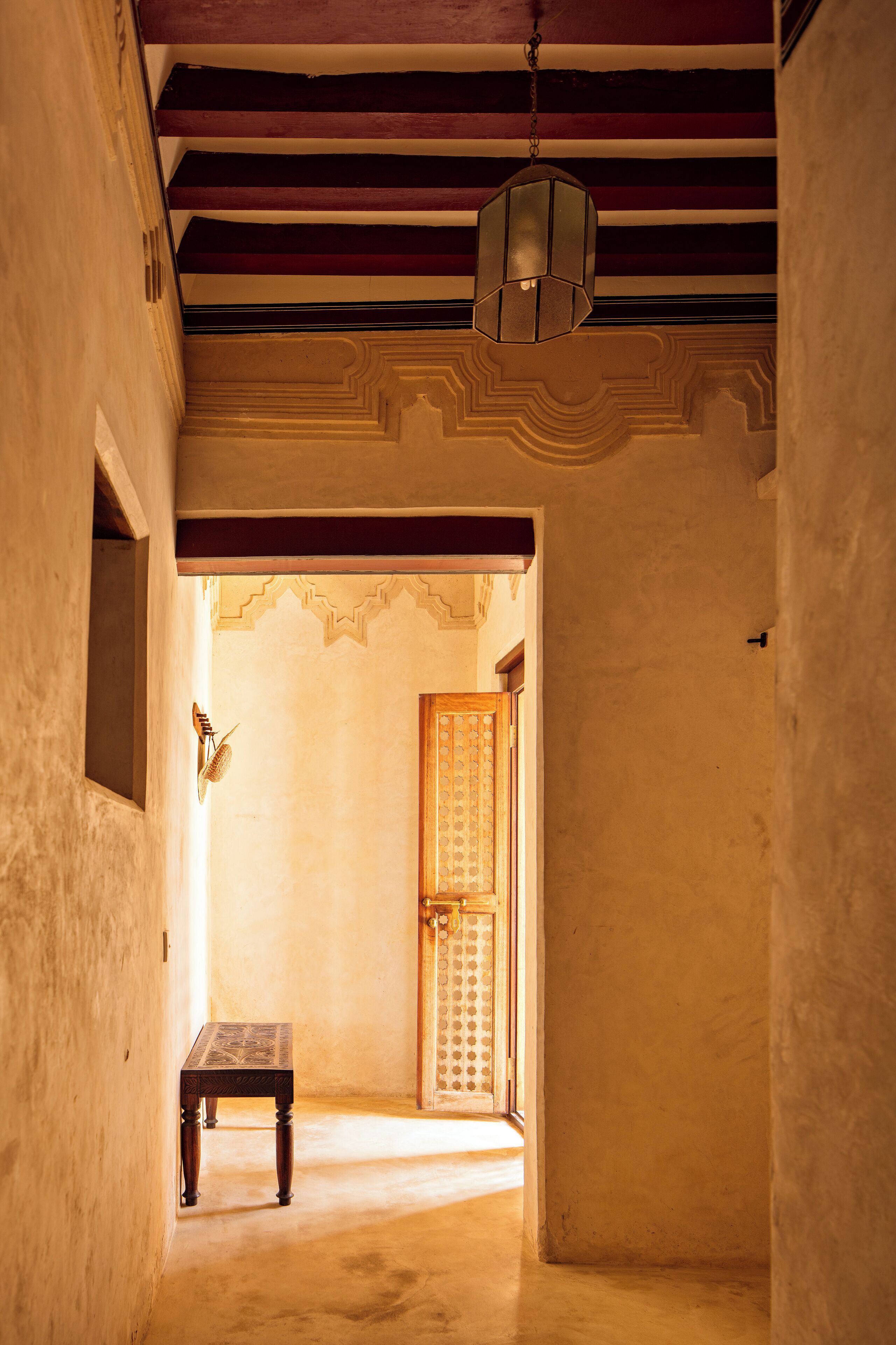 A narrow hallway with a wooden bench, a hanging lantern, and a wooden door with intricate patterns.