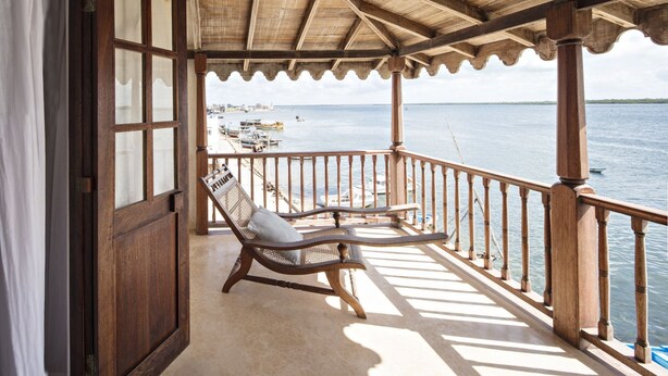 A wooden balcony with a thatched roof overlooking a calm sea. There are two wicker chairs with cushions.