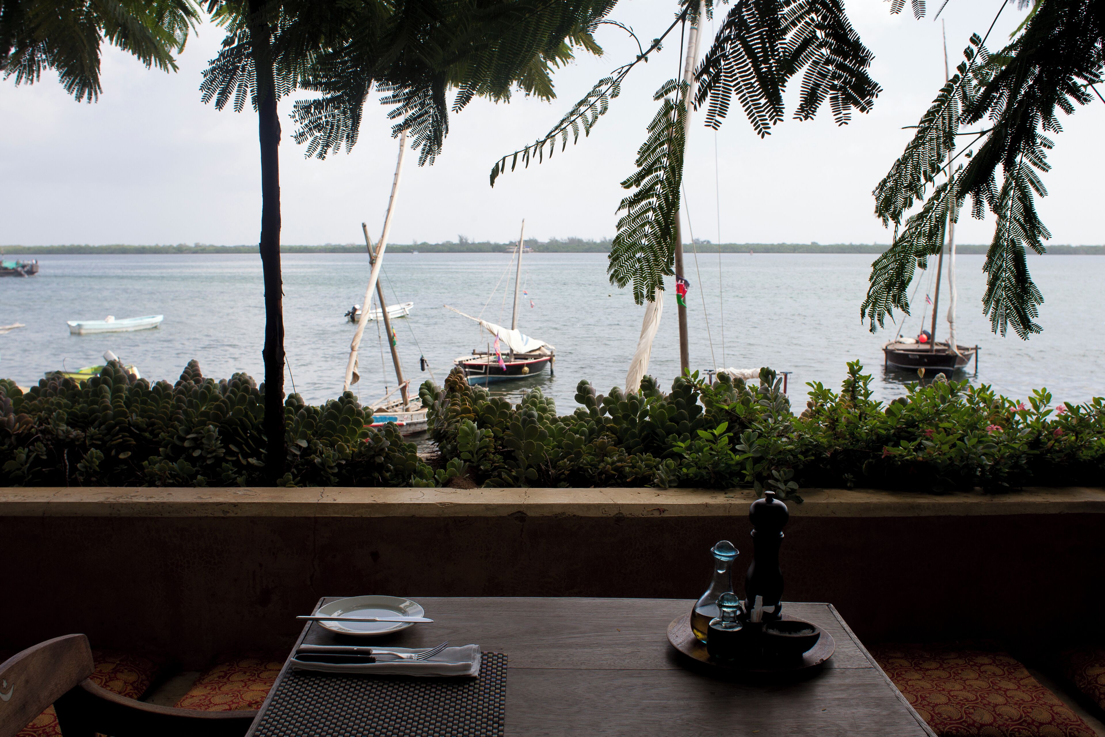 Une table avec un set de table, une bouteille et une tasse, donnant sur une étendue d’eau calme où flottent des bateaux.