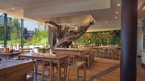A modern restaurant interior with wooden tables and chairs, a central island with a marble countertop, and a large tree sculpture. - Center Parcs Villages Nature Paris (Villeneuve-le-Comte)