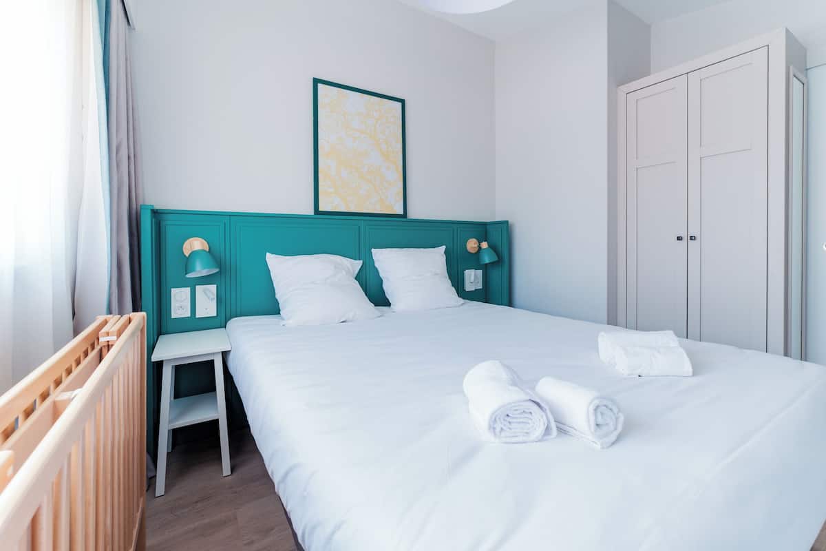 A neatly made bed with white linens and rolled towels, a teal headboard, a small bedside table with a lamp, and a framed picture on the wall.
