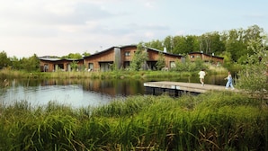 A modern building with a wooden exterior near a body of water, featuring a dock and lush greenery. - Center Parcs Villages Nature Paris (Villeneuve-le-Comte)