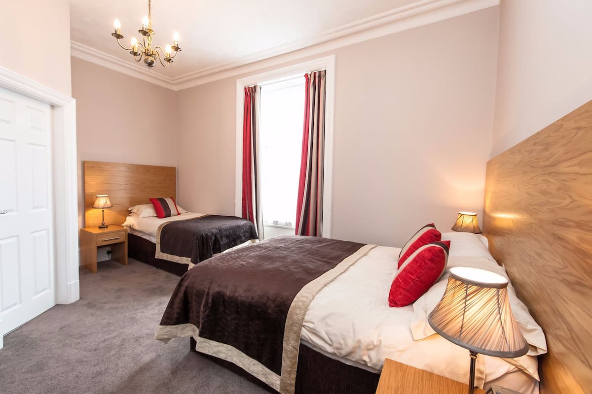 A hotel room with two beds, a chandelier, a window with curtains, and a bedside lamp.