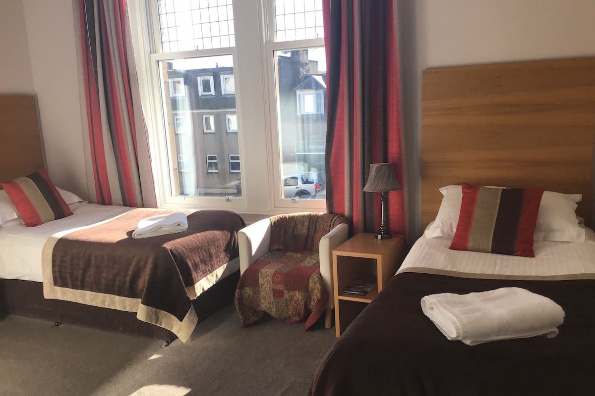 A hotel room with two beds, a small table, a chair, and a window with curtains.