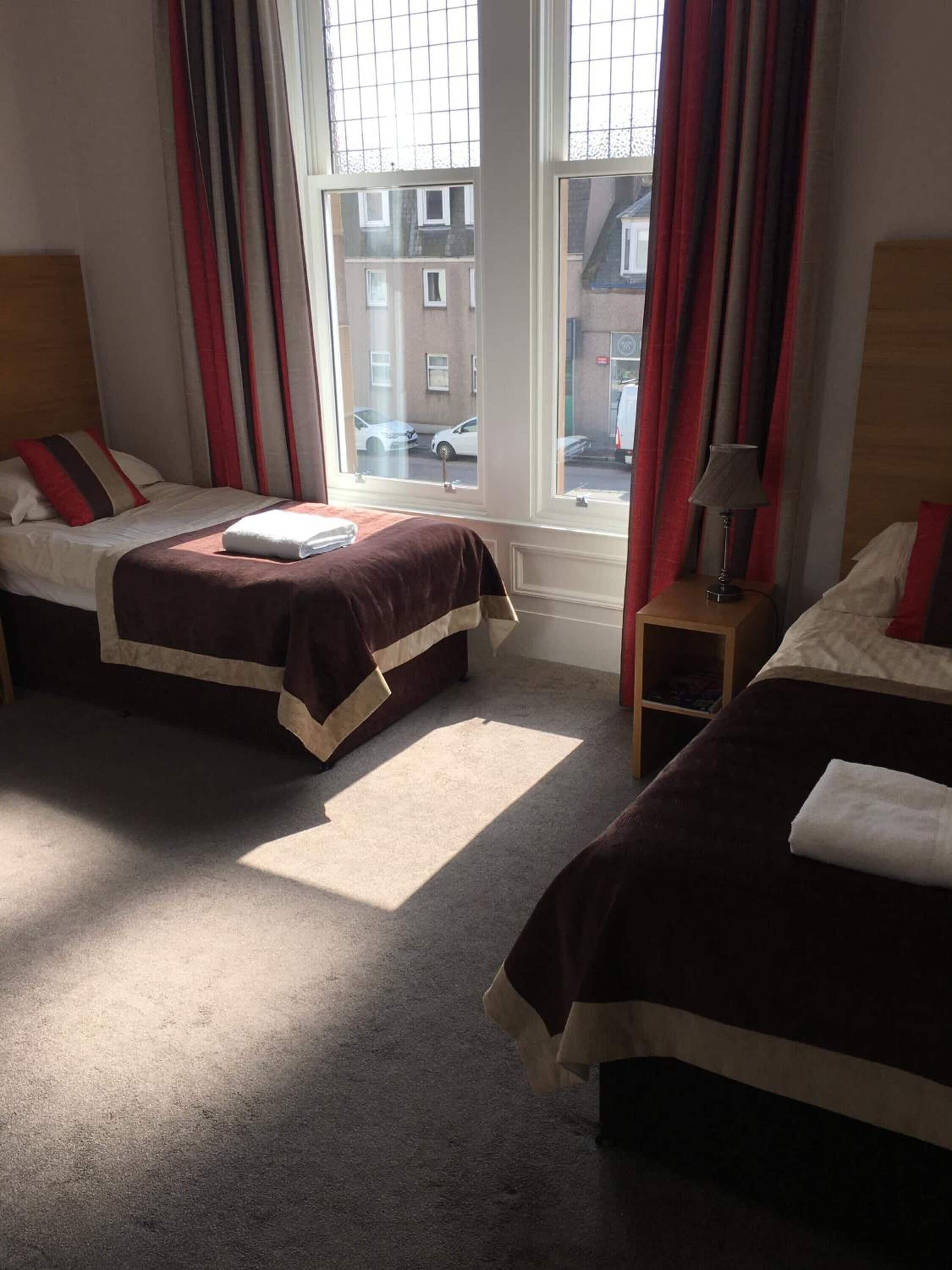 A hotel room with two beds, a window with curtains, a lamp on a nightstand, and a view of the street outside.