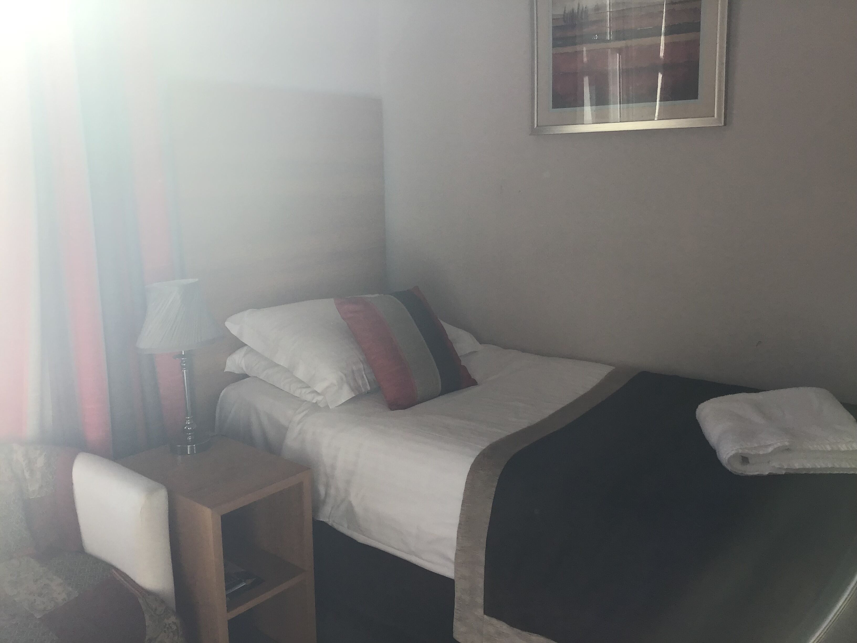 A hotel room with a bed, a bedside table with a lamp, a chair, and a framed picture on the wall.