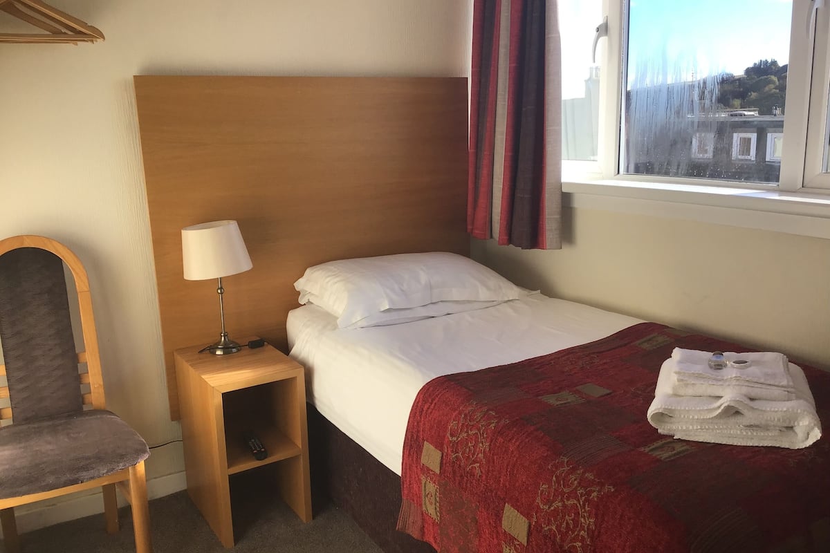 A hotel room with a single bed, a wooden nightstand with a lamp, a chair, and a window with curtains.