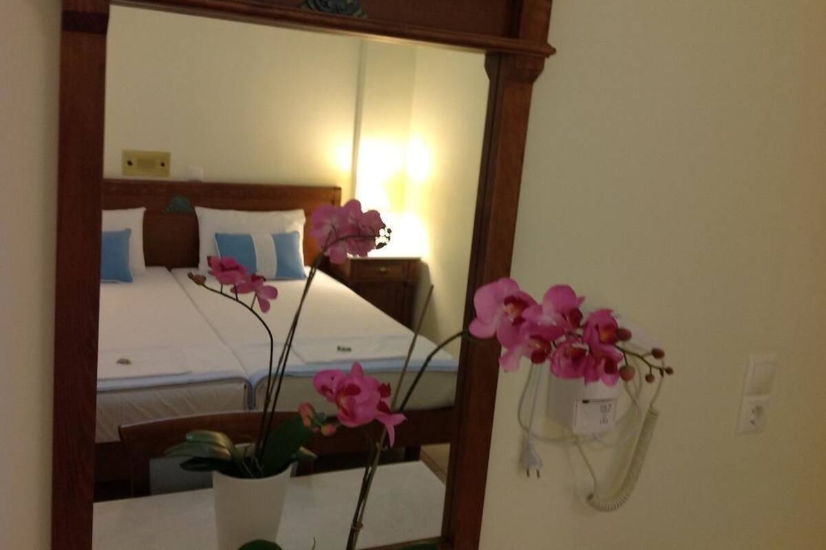 A hotel room with a bed, a nightstand, a mirror, and a vase with pink flowers.