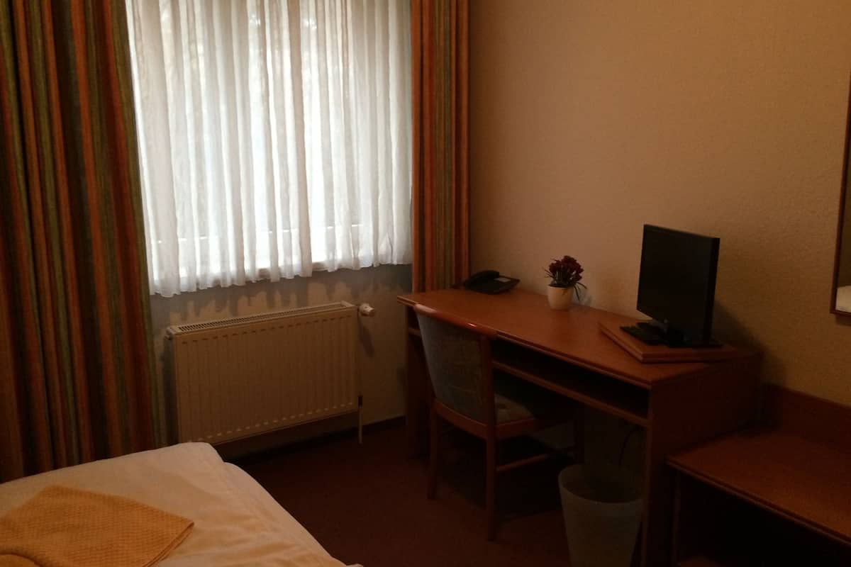 A hotel room with a bed, a desk with a chair, a radiator, a window with curtains, and a small trash bin.