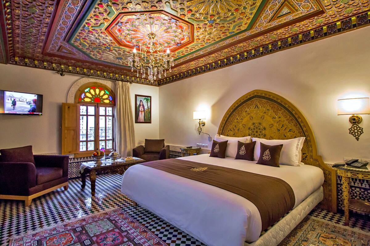 A hotel room with a large bed, ornate ceiling, and a television on the wall.