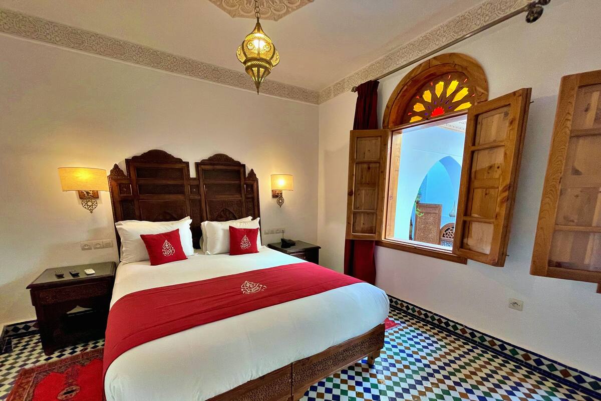 A hotel room with a large bed, two bedside lamps, a decorative ceiling light, a checkered floor, and a window with an open shutter.