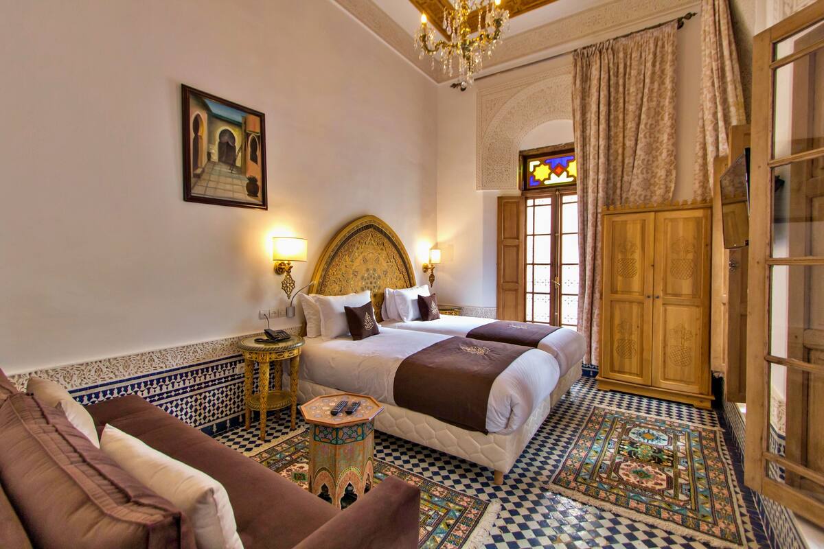 A traditional Moroccan room with a large bed, a sofa, a small table with a lamp, and a decorative rug.