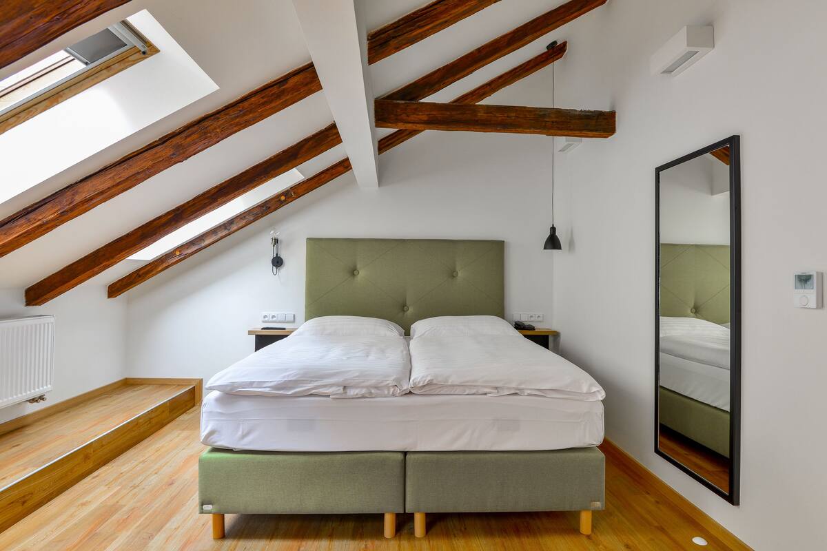 A bedroom with a sloped ceiling, wooden beams, a green upholstered bed, a bedside table, a wall-mounted mirror, and a radiator.