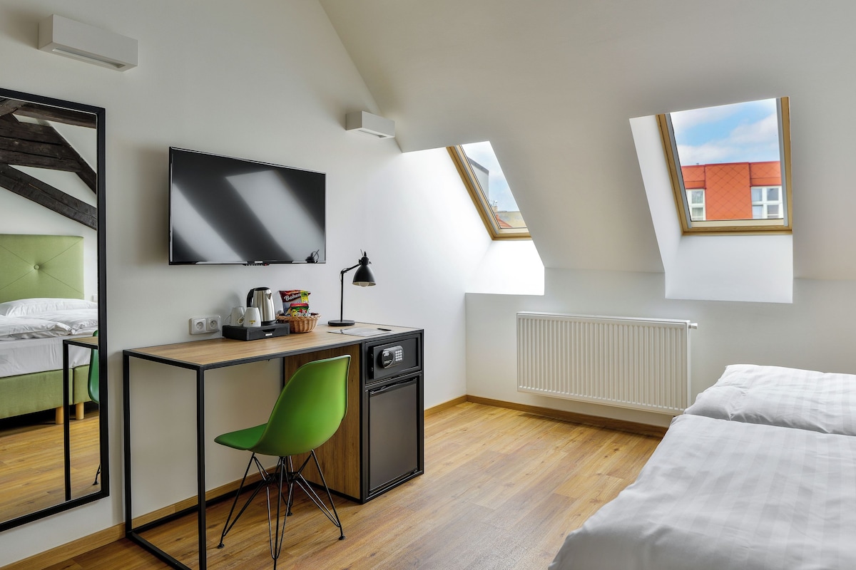 A modern hotel room with a bed, a desk with a chair, a television, a radiator, and a window with a view of a building.