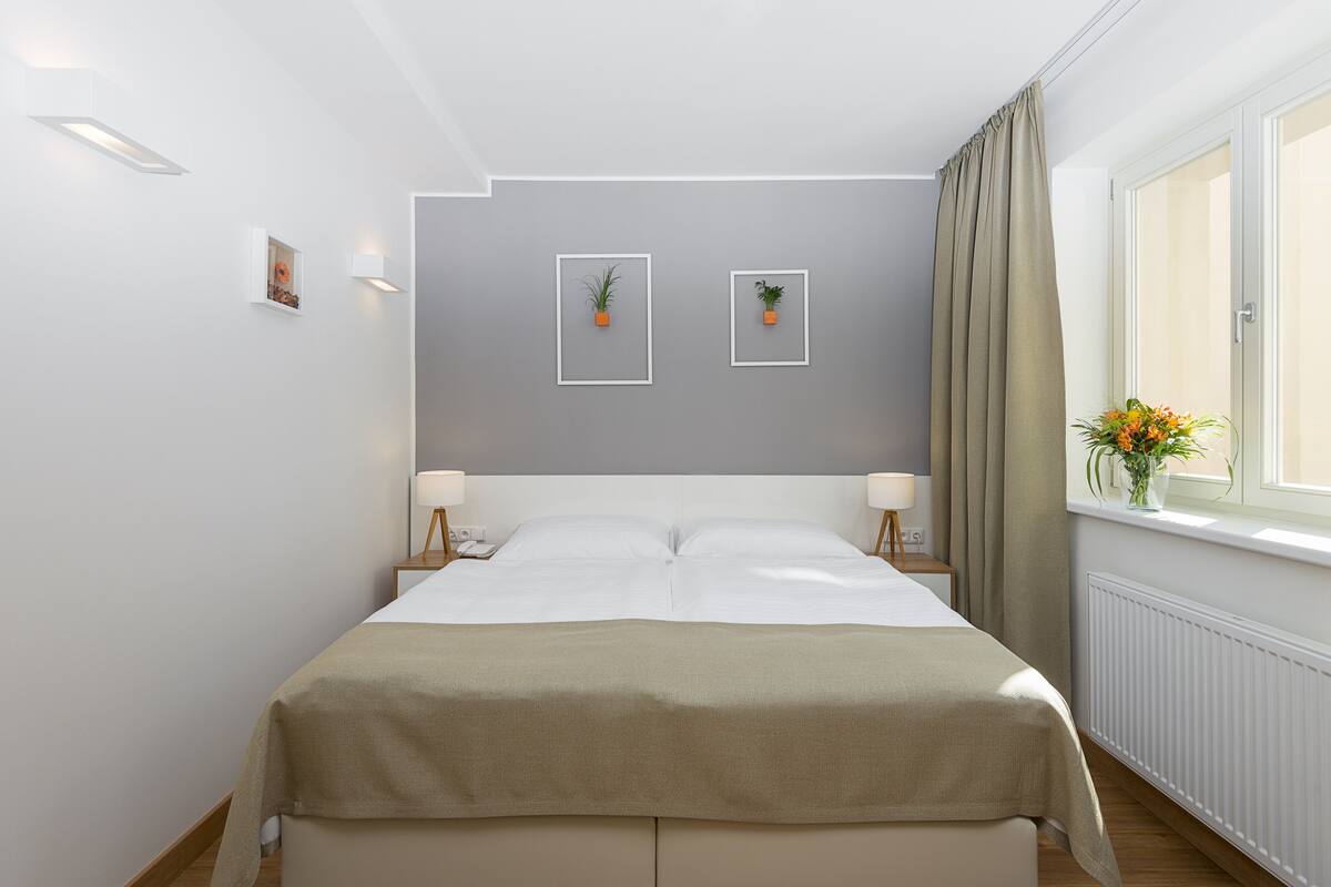A neatly arranged bedroom with a bed, bedside tables, a window with curtains, and wall-mounted plants.