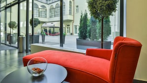 A modern hotel lobby with a red sofa, a glass table with a decorative bowl, and large windows offering a view of the building's interior and a tree outside. - Hotel Garden Court (Prague)