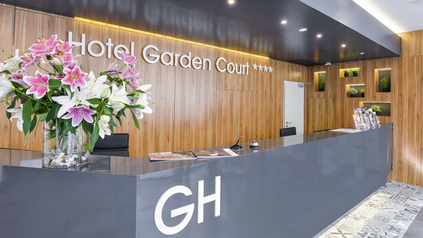 A hotel reception desk with a wooden panel wall, a large floral arrangement, and a sign reading "Hotel Garden Court". - Hotel Garden Court (Prague)