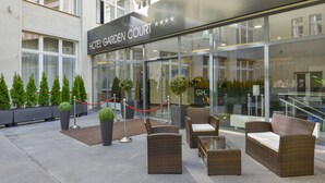 A hotel entrance with a glass facade, outdoor seating area with wicker furniture, and neatly arranged potted plants. - Hotel Garden Court (Prague)