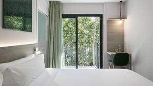 Habitación de hotel con cama, escritorio con silla, balcón y vistas a árboles, además de una lámpara colgante.