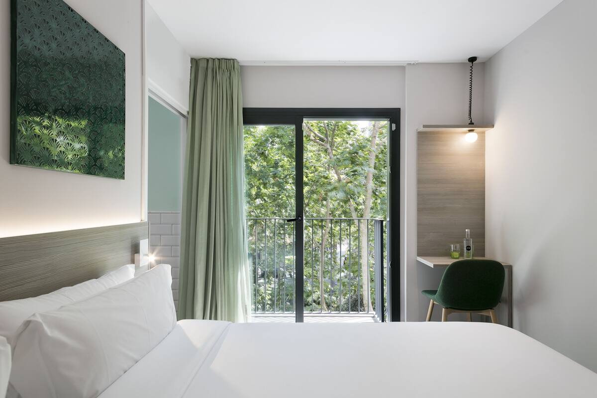 A hotel room with a bed, a desk with a chair, a balcony with a view of trees, and a hanging light fixture.