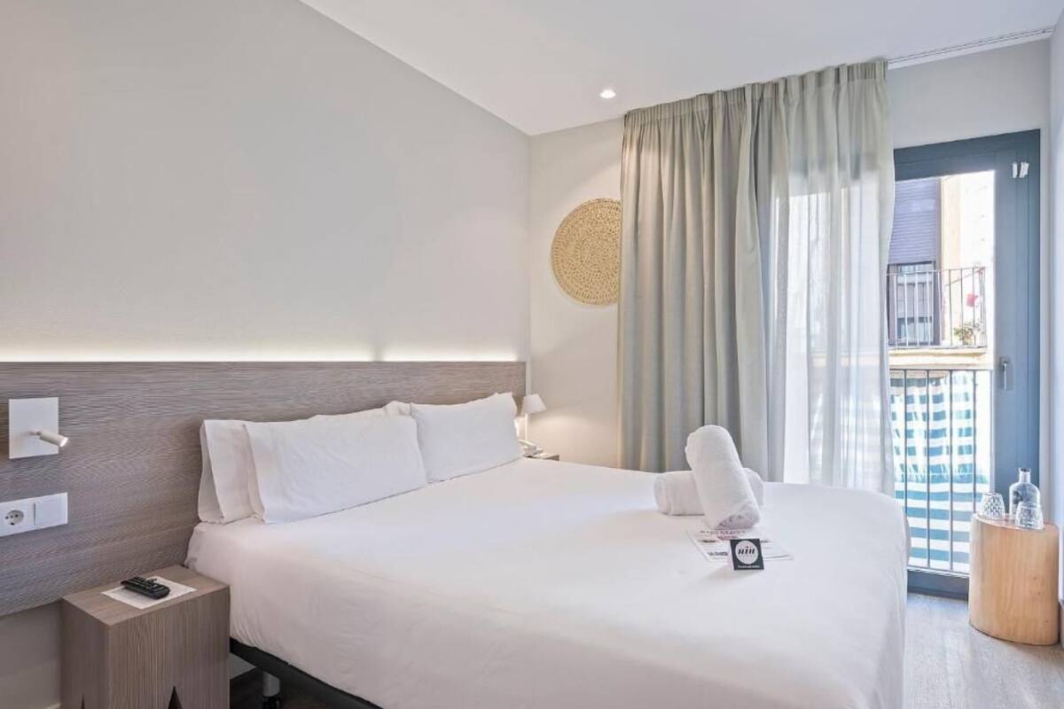 A hotel room with a large bed, a bedside table, a nightstand, a round wall decoration, a window with curtains, and a door leading to an outdoor balcony.