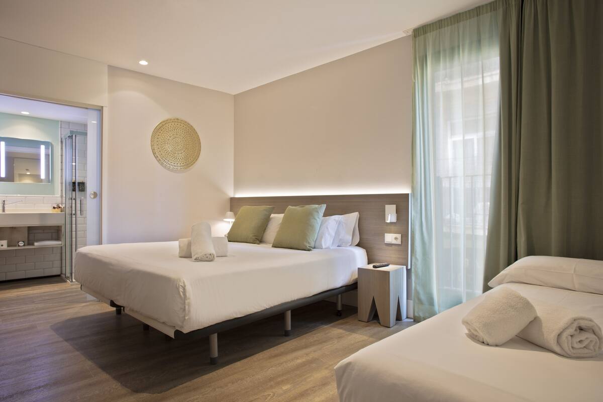 A modern hotel room with a large bed, bedside table, and a view of a cityscape through the window.