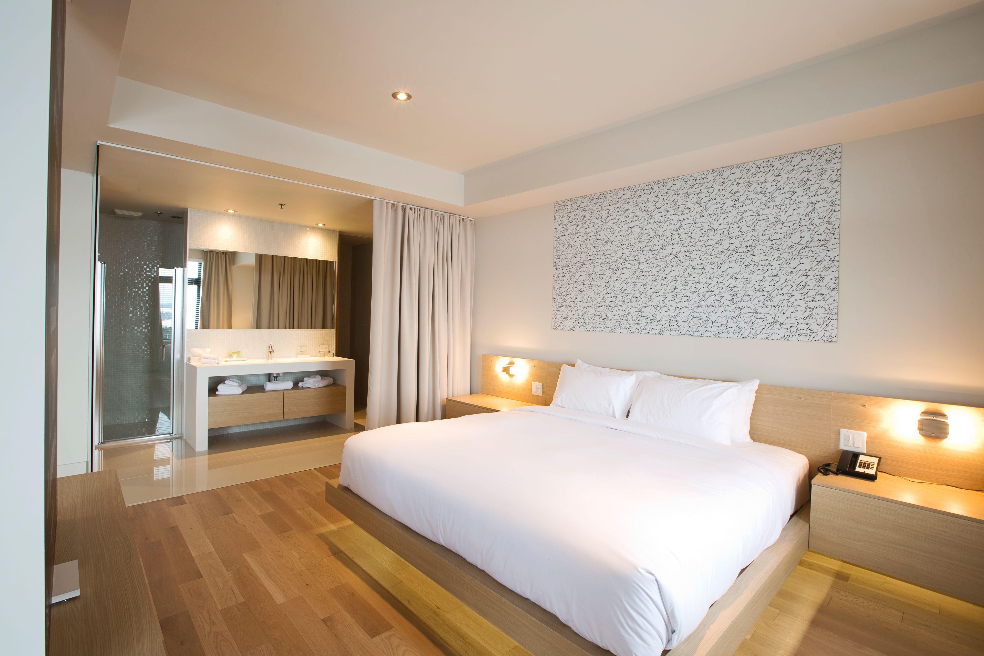 A modern hotel room with a large bed, a bathroom with a glass shower, and a wall-mounted light fixture.