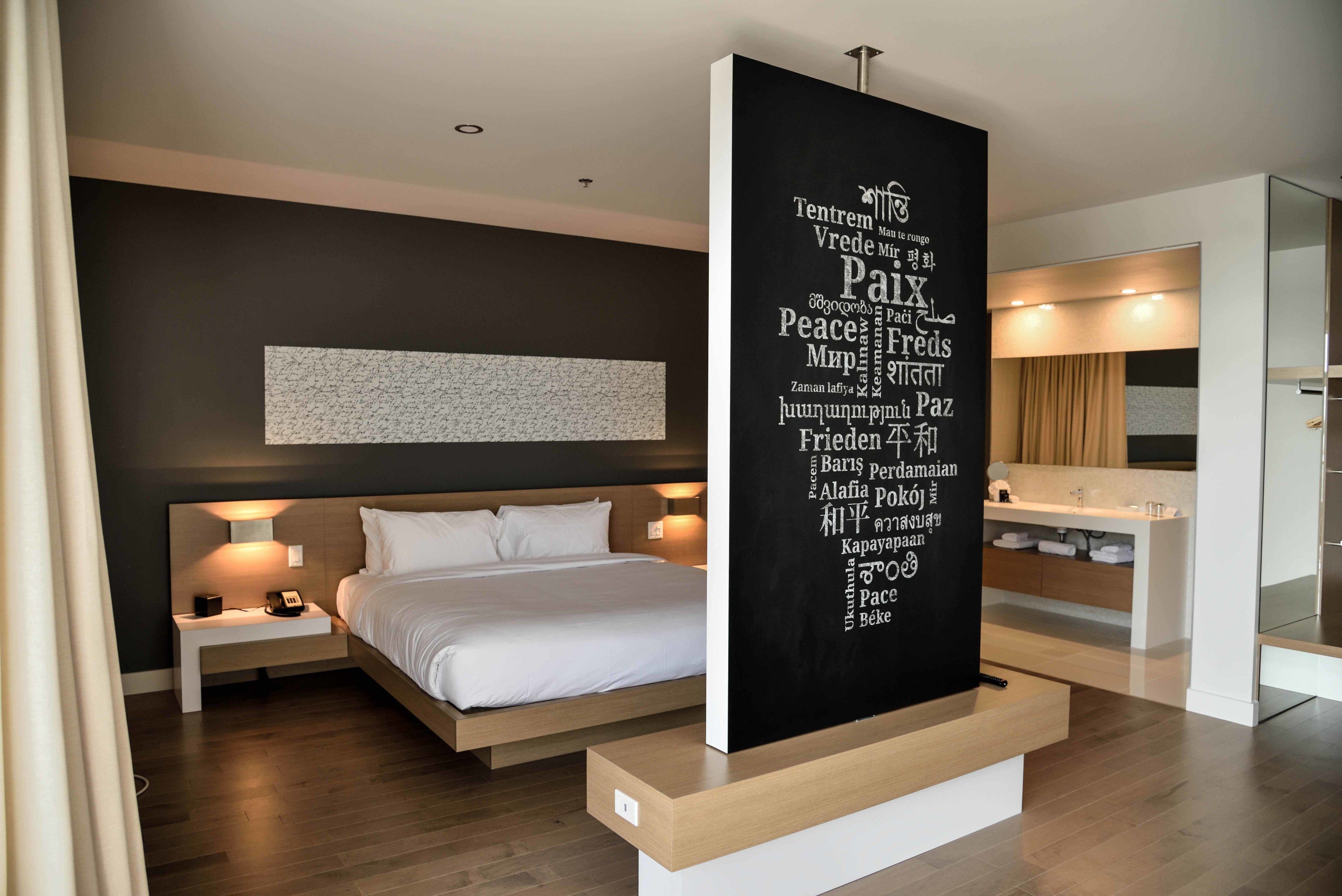 Een moderne hotelkamer met een groot bed, nachtkastjes, een kunstwerk aan de muur en een badkamer met wastafel en spiegel.