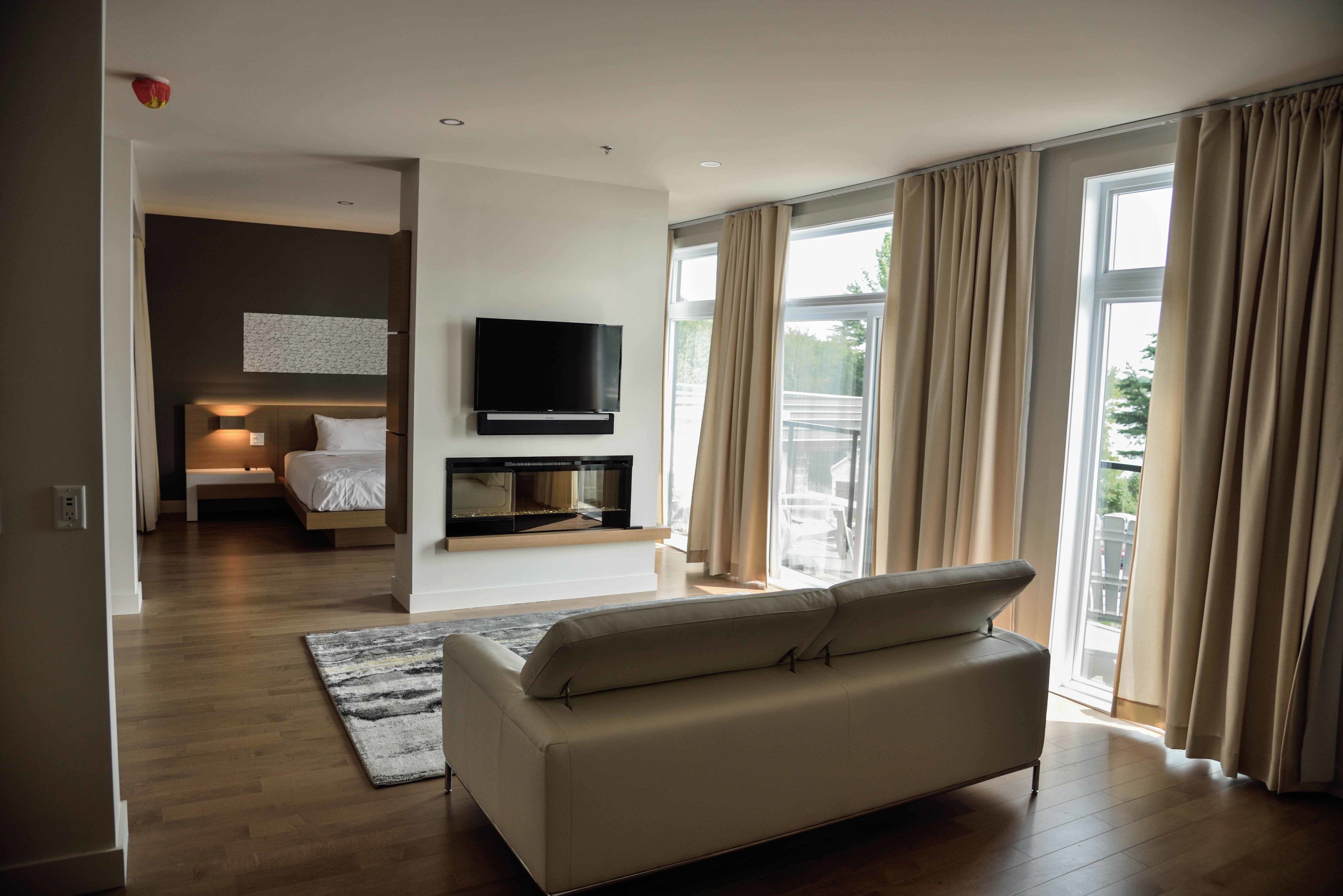 A modern living room with a sofa, a flat-screen TV mounted on the wall, and a large window with curtains.