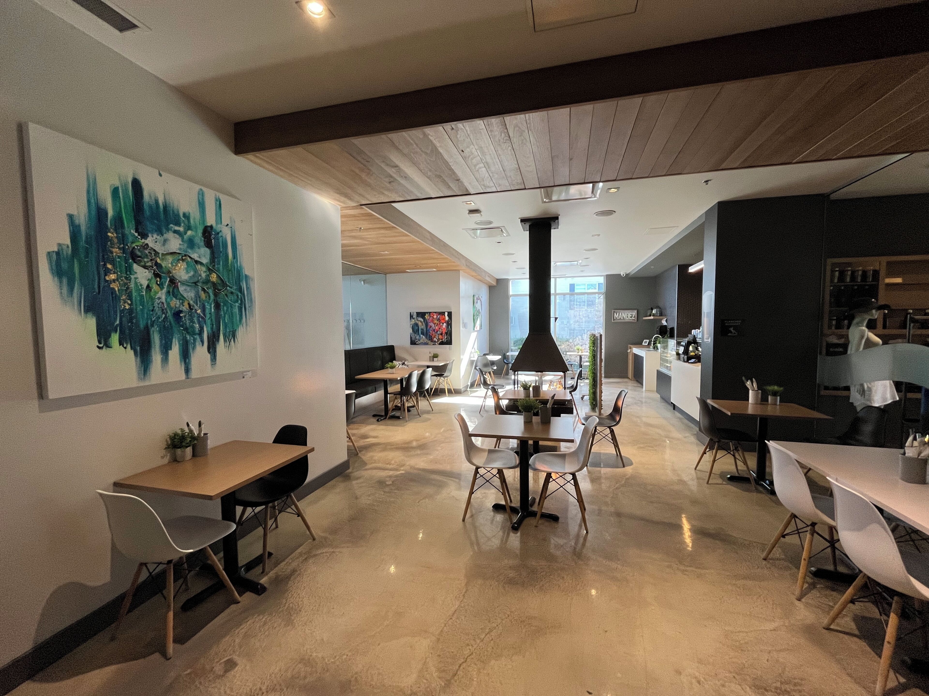 Un comedor moderno con mesas y sillas, un cuadro abstracto grande en la pared y una cocina al fondo.