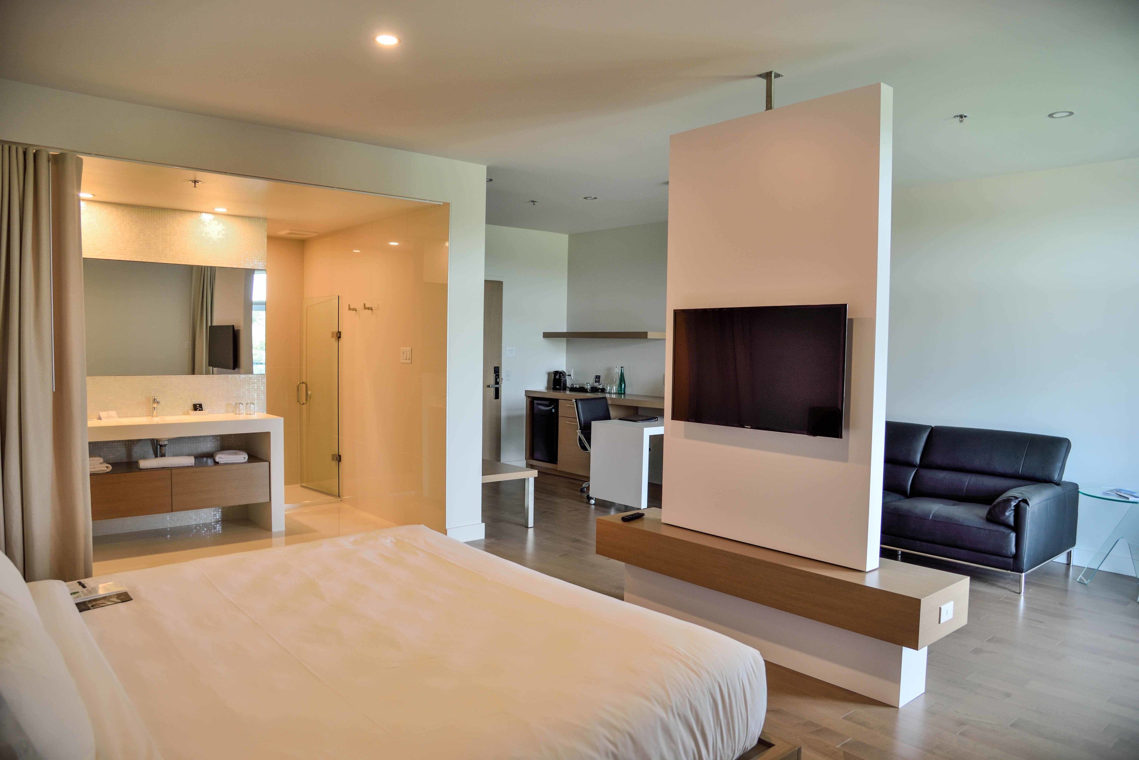 A modern hotel room with a large bed, a TV mounted on a wall, a separate seating area with a sofa, and a kitchenette in the background.