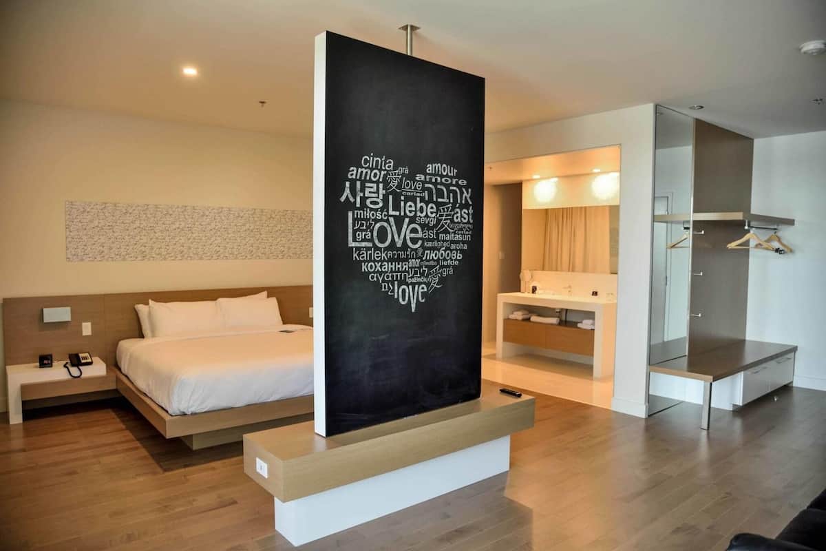 A modern hotel room with a bed, bedside table, a large wall art piece, a built-in bench, a mirror, and a wardrobe.