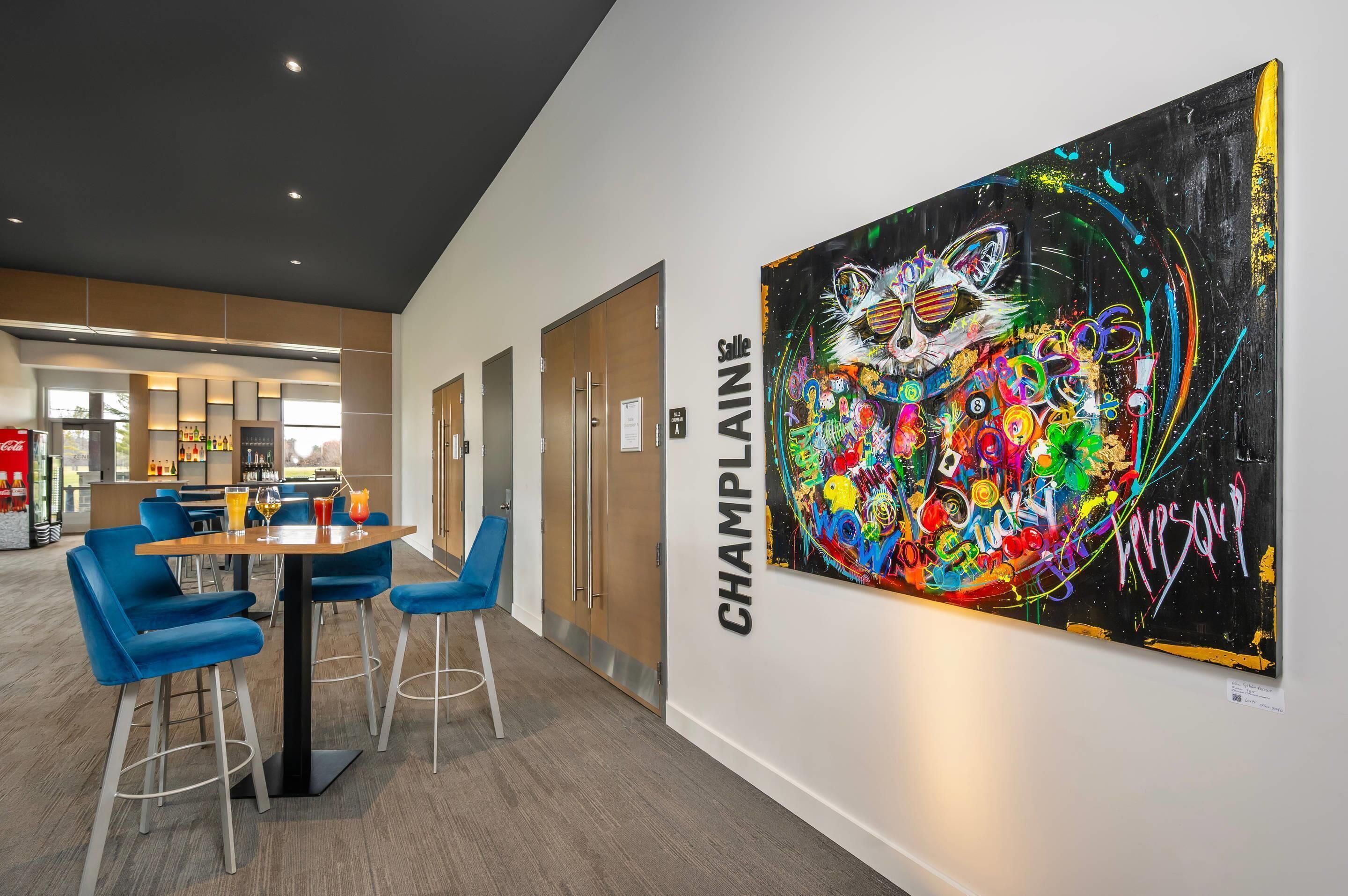 A modern interior with a dining area featuring blue chairs and a table, a large colorful abstract painting on the wall, and a sign reading 'CHAMPLAIN'.