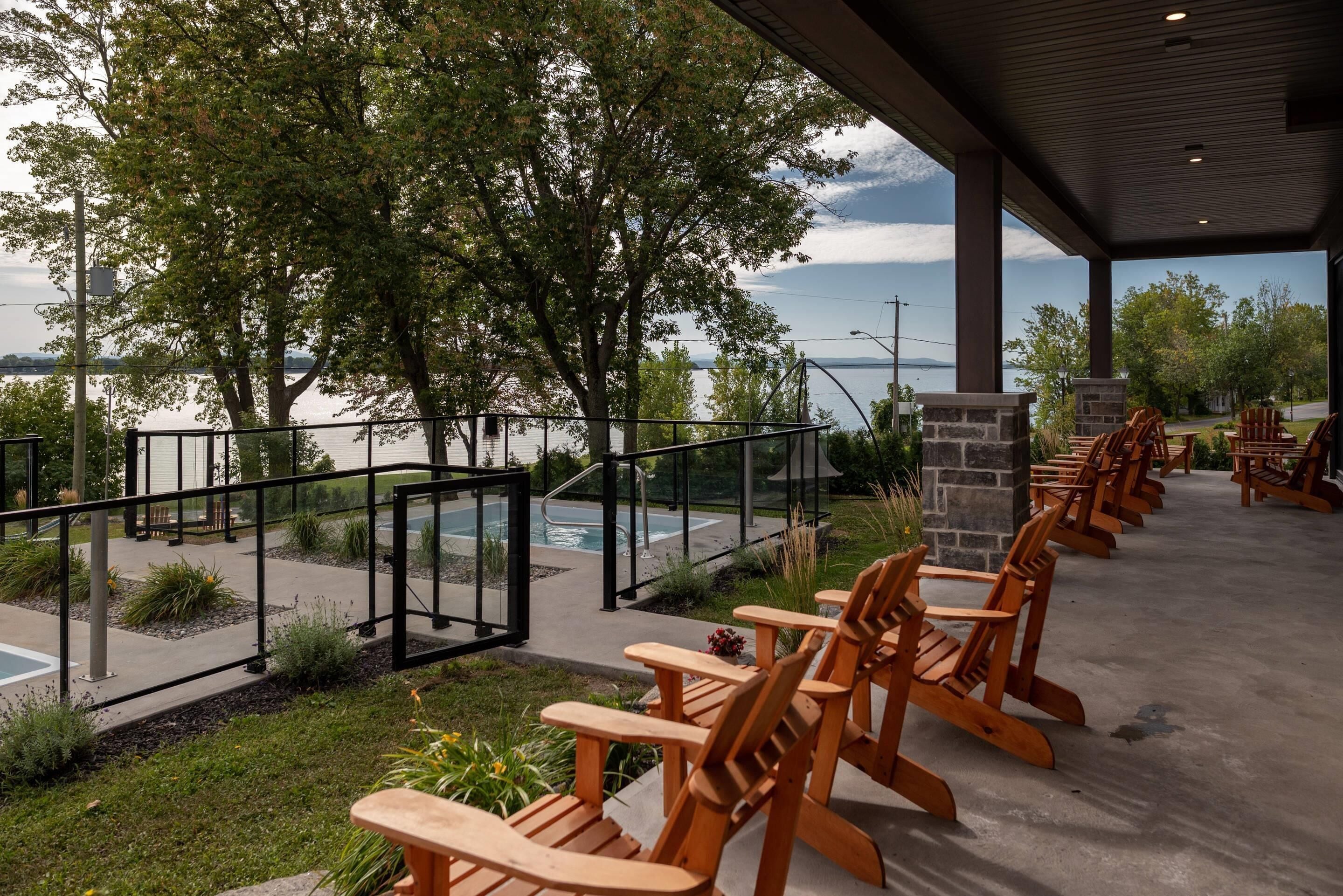 A covered outdoor seating area with wooden chairs and tables, overlooking a body of water with trees and a clear sky.