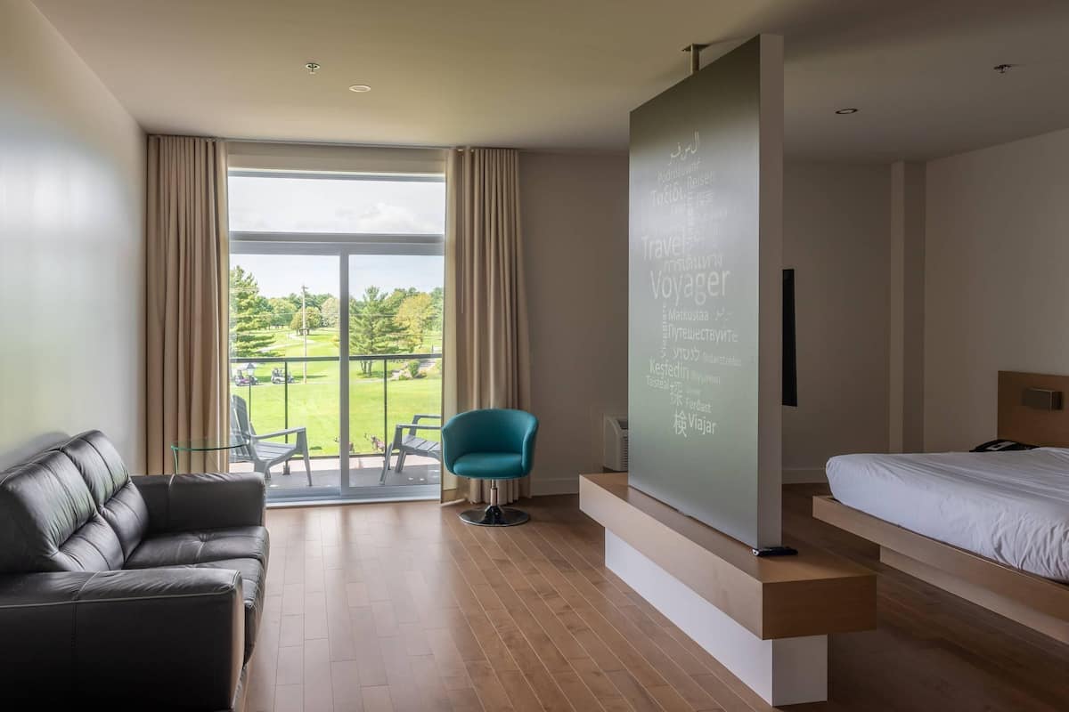 A modern hotel room with a large window, a sofa, a chair, a bed, and a projection screen.