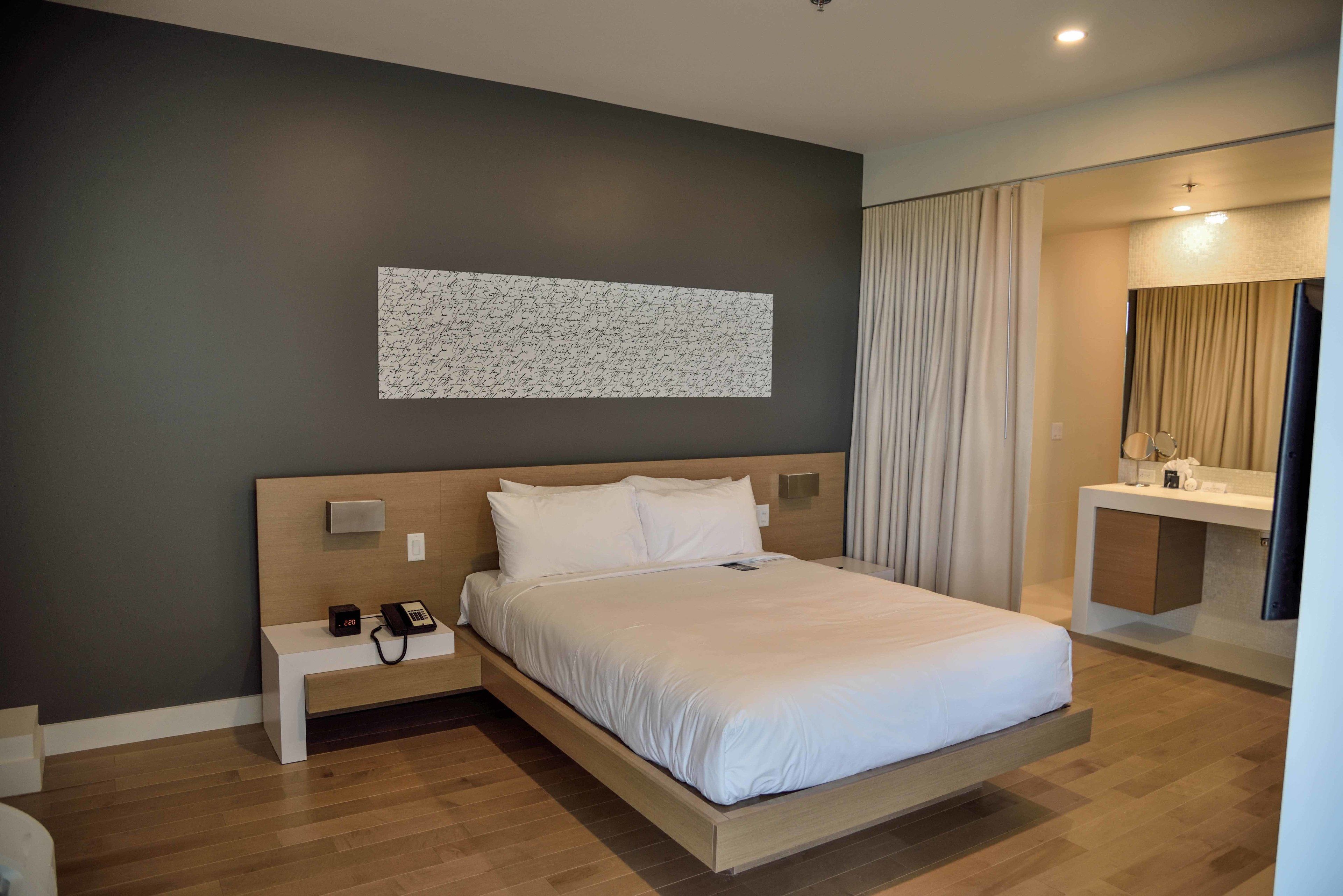 A modern hotel room with a large bed, bedside table with a phone, and a small bedside table with a lamp.